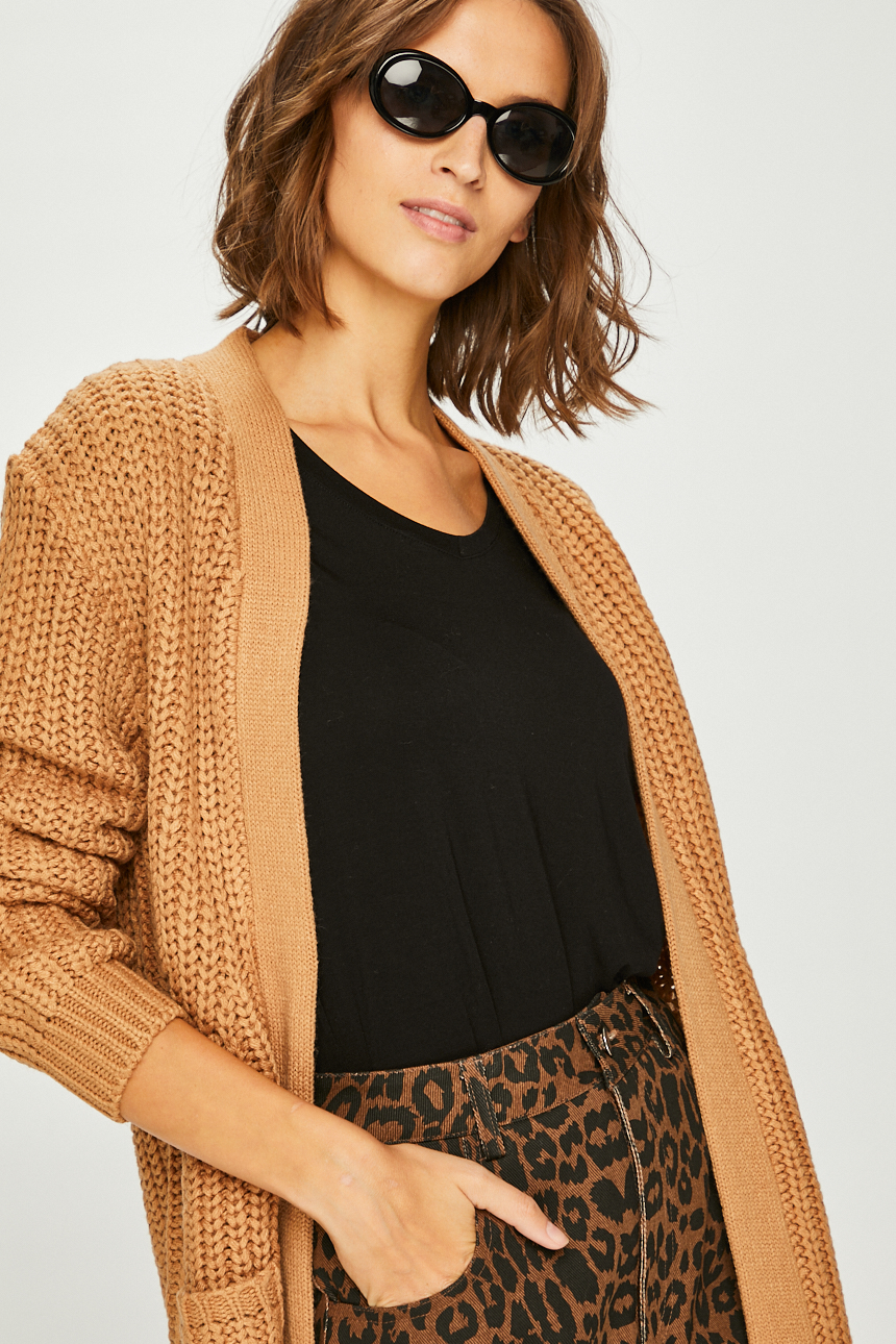 Kobieta w okularach przeciwsłonecznych, ubrana w czarny top i rozpinany, dziergany, brązowy kardigan, pozująca na jasnym tle.