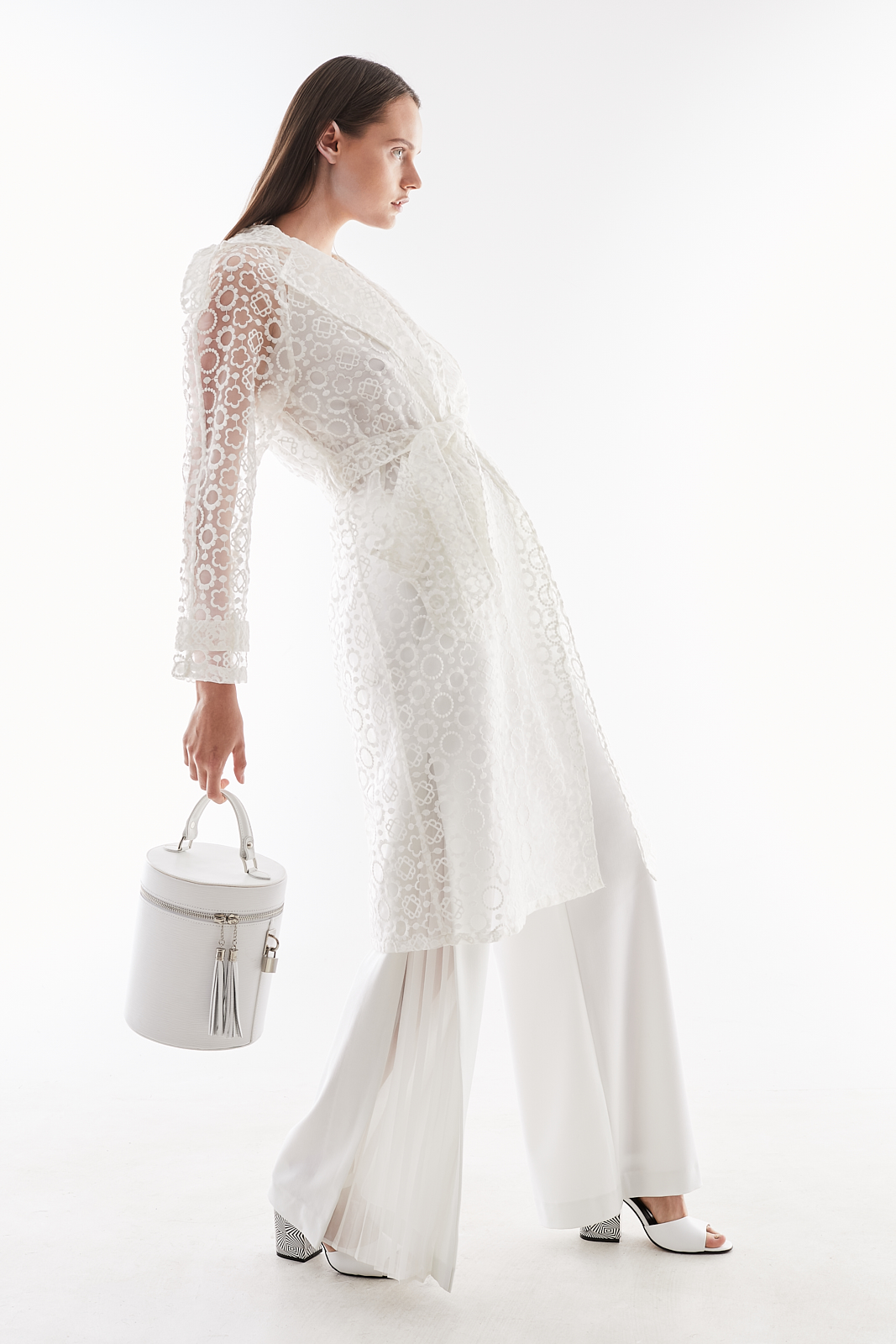 Modelka w białym koronkowym płaszczu, szerokich spodniach i butach na obcasie, trzymająca białą okrągłą torebkę z frędzlami na tle jasnego studia.
