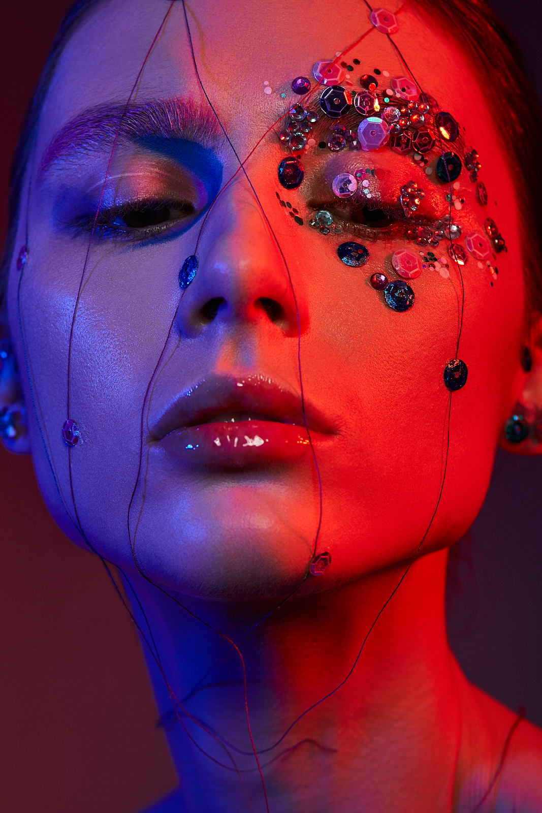 Portret kobiety z artystycznym makijażem: kolorowe cekiny wokół oka i cienkie nitki zdobiące twarz, oświetlone niebieskim i czerwonym światłem.