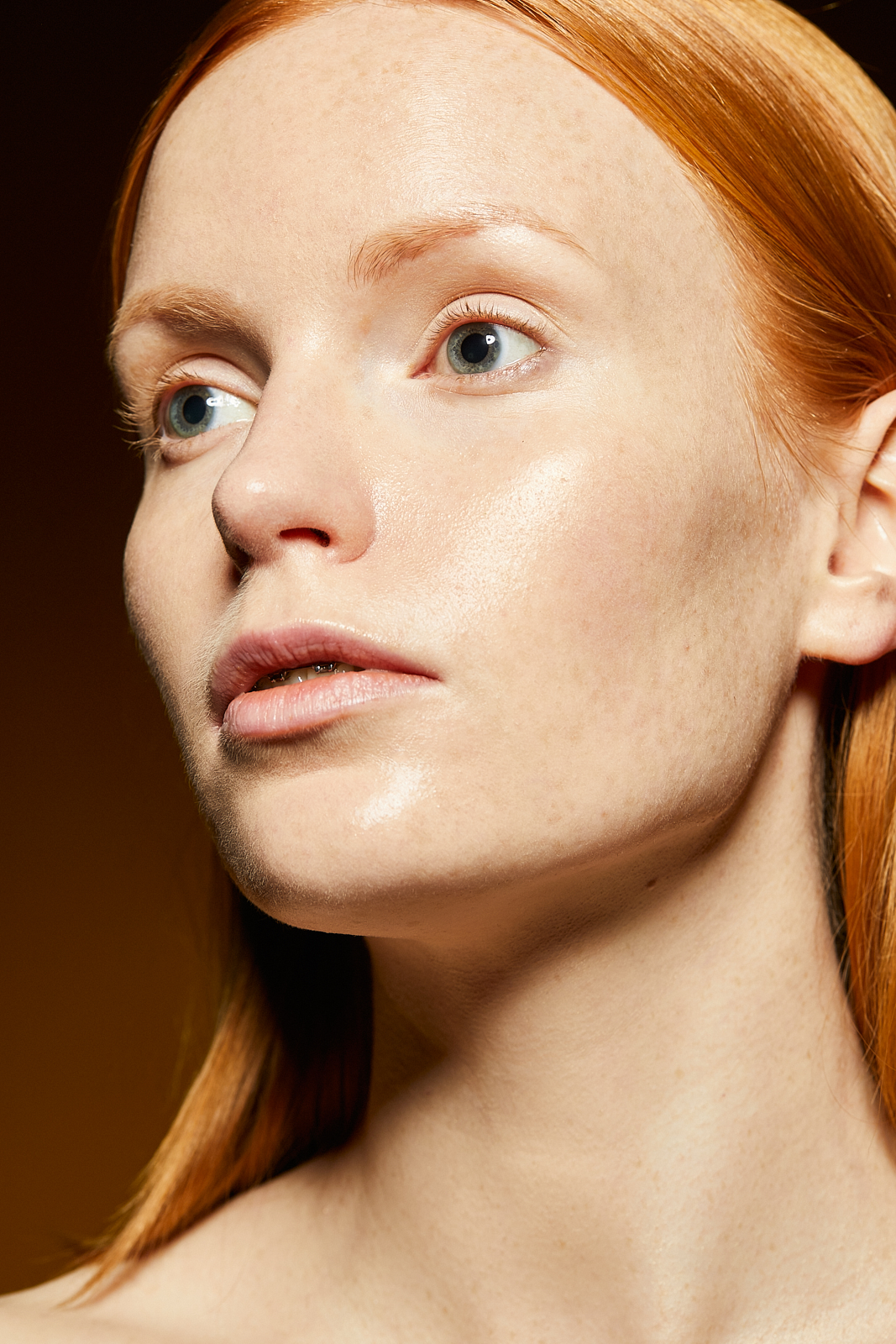 Portret kobiety o rudych włosach, z jasną cerą i piegami, z delikatnym makijażem i błyszczącą skórą, skierowana wzrokiem w lewo, na ciemnym tle.