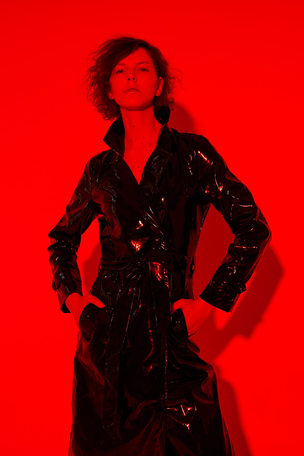 Kobieta w błyszczącym, czarnym płaszczu z paskiem, stojąca na tle intensywnej czerwieni, ręce w kieszeniach, ostre światło podkreśla fakturę materiału.