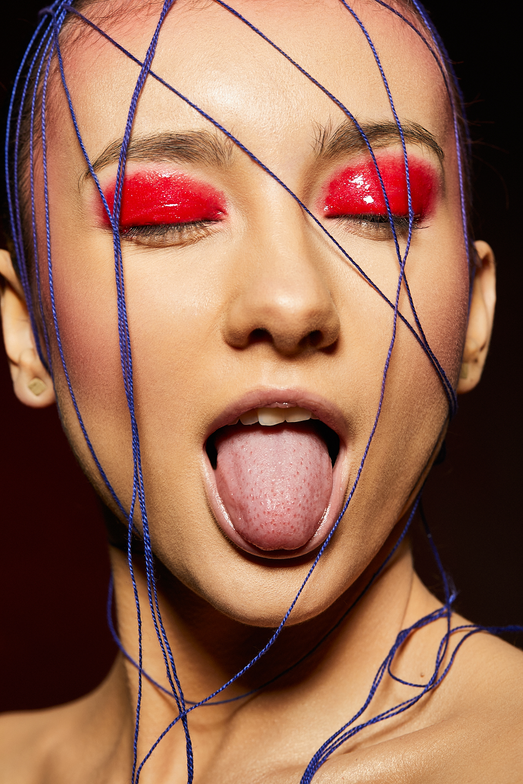 Portret kobiety z otwartymi ustami, wyciągniętym językiem, intensywnym czerwonym makijażem oczu i niebieskimi sznurkami oplatającymi twarz.