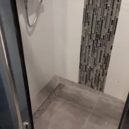 Nowoczesna kabina prysznicowa z odpływem liniowym w podłodze i dekoracyjną mozaiką na ścianie, widok z poziomu podłogi, szare płytki podłogowe.