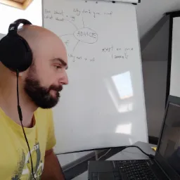 Mężczyzna z brodą i słuchawkami na uszach, prowadzi lekcję angielskiego online, widoczny laptop z odbiciem osoby, za nim tablica z napisem 'ADVICE' i przykładami zdań.