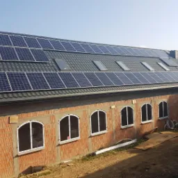 Niedokończony budynek z cegły z zamontowanymi panelami słonecznymi na grafitowym dachu z oknami dachowymi, widoczne niedokończone otwory okienne z białymi ramami i palety w tle.