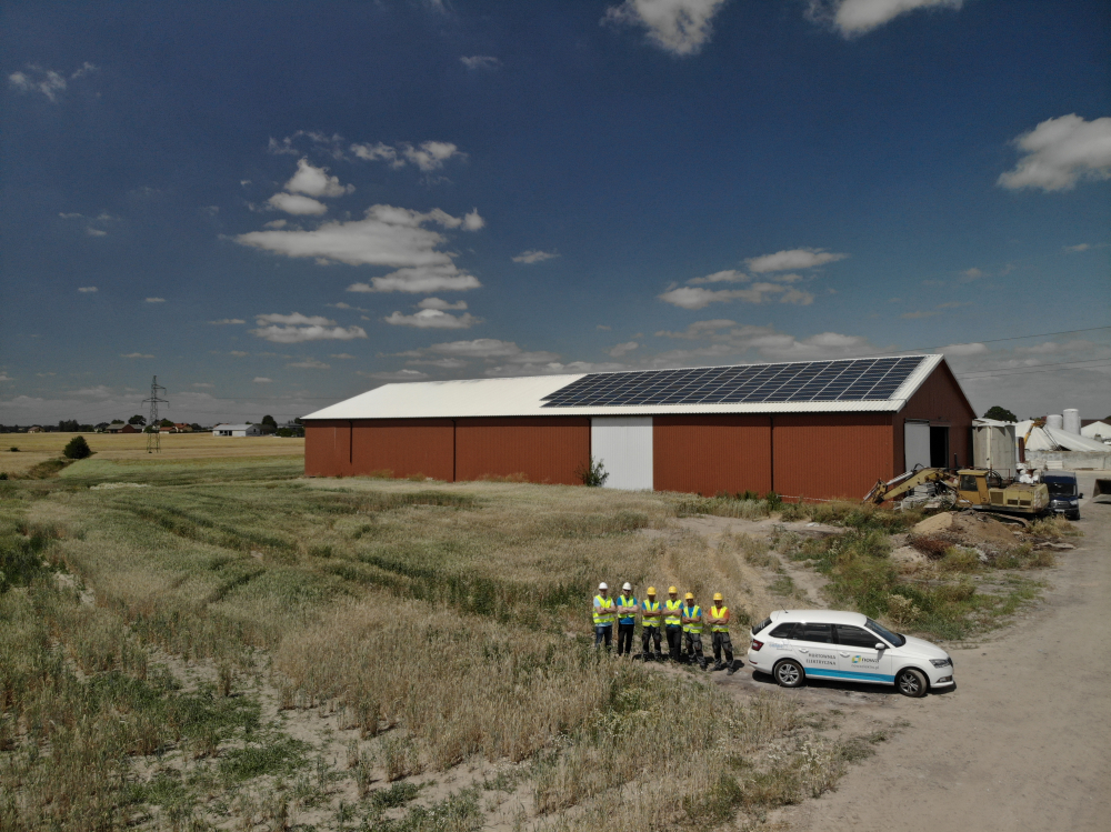 Szeroki kadr na budynek gospodarczy z panelami słonecznymi na dachu, na tle pola i błękitnego nieba z chmurami; grupa pracowników w kamizelkach odblaskowych i kaskach stoi przed budynkiem obok...