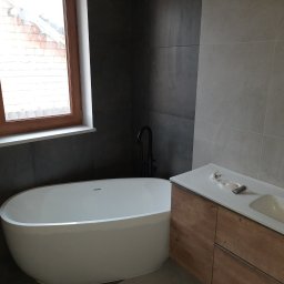 SUPRA PRZEDSIĘBIORSTWO BUDOWLANE - Nowoczesna łazienka z wolnostojącą białą wanną, szarą glazurą na ścianach, drewnianą szafką pod umywalką z czarną armaturą oraz oknem z widokiem na ceglany budynek.
