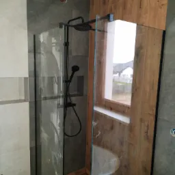 Nowoczesna kabina prysznicowa z czarną armaturą, wykończona płytkami imitującymi drewno i szarym gresem, z widokiem na zabudowę jednorodzinną przez okno.