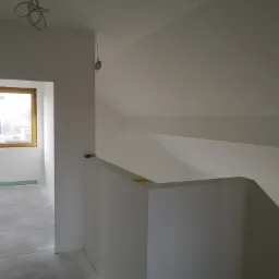 Poddasze po pracach wykończeniowych: gładkie, białe ściany i sufit, widoczne okno z drewnianą ramą oraz wystające przewody elektryczne.