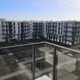 SUPRA PRZEDSIĘBIORSTWO BUDOWLANE - Widok z balkonu na plac budowy nowego osiedla z pracownikami w żółtych kamizelkach sadzącymi drzewa i krzewy w betonowych donicach.