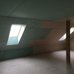 SUPRA PRZEDSIĘBIORSTWO BUDOWLANE - Poddasze w trakcie wykańczania: widoczne skośne ściany z zamontowaną częściowo zieloną płytą, okna dachowe w ścianach oraz drewniany słup podtrzymujący konstrukcję.