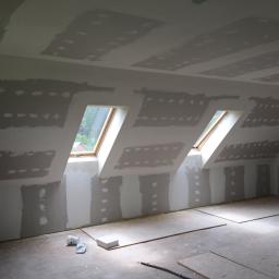 SUPRA PRZEDSIĘBIORSTWO BUDOWLANE - Poddasze z dwoma oknami dachowymi w trakcie szpachlowania płyt kartonowo-gipsowych, widoczne łączenia płyt i częściowo zagruntowane powierzchnie, na podłodze resztki materiałów budowlanych.