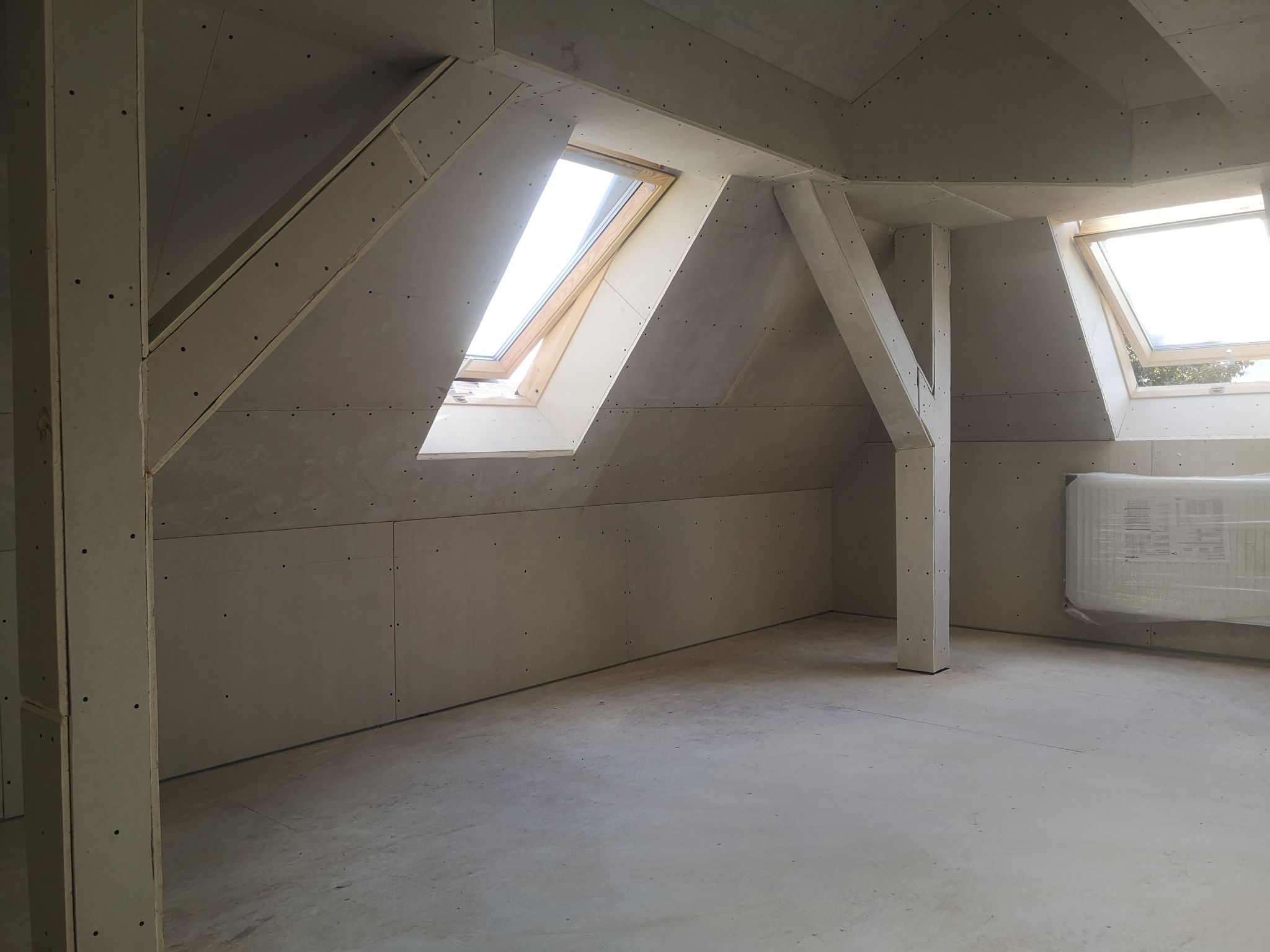 Poddasze w trakcie wykańczania: zamontowane płyty kartonowo-gipsowe, widoczne drewniane okna dachowe i elementy konstrukcyjne, przygotowane do dalszych prac wykończeniowych.