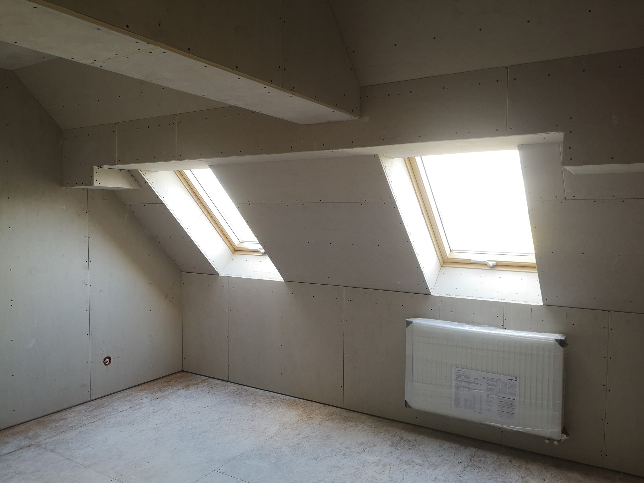 Poddasze z oknami dachowymi i grzejnikiem w folii, ściany i sufit wykończone płytami karton gips, widoczne łączenia płyt.