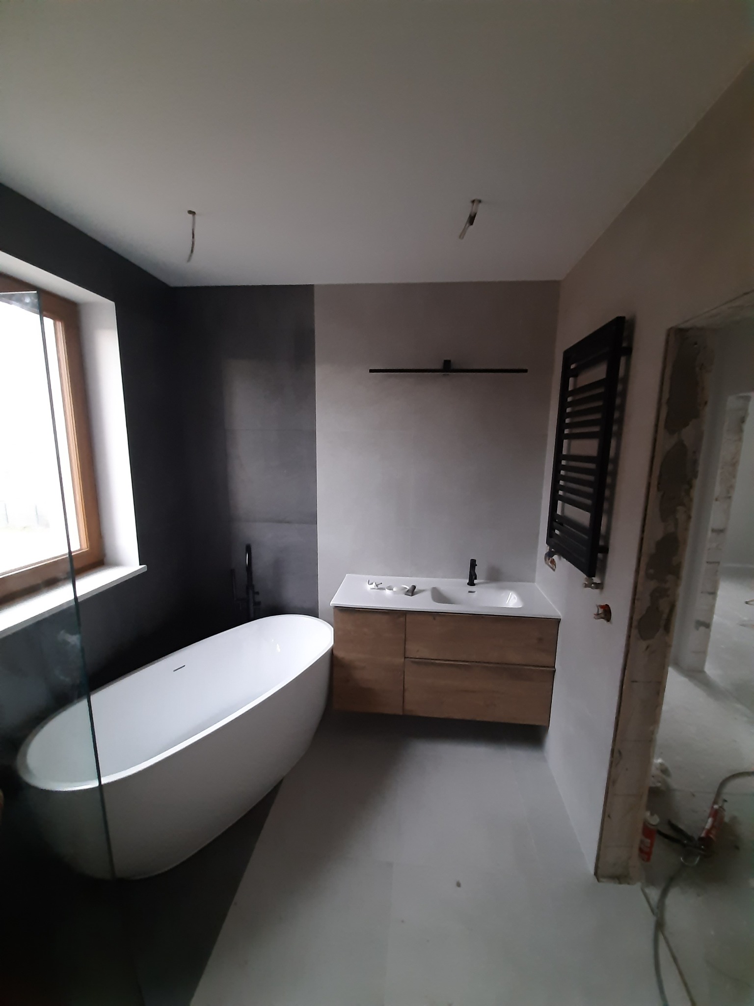Nowoczesna łazienka w trakcie wykańczania: wolnostojąca wanna przy oknie, drewniana szafka z umywalką i czarny grzejnik drabinkowy na ścianie. Widoczne niedokończone prace przy ościeżnicy drzwi.