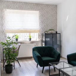 Między Myślami - Jasne wnętrze pokoju z zielonymi fotelami, stolikiem kawowym, biurkiem z krzesłem i roślinami doniczkowymi przy oknie z roletą rzymską.