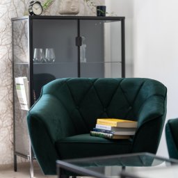 Między Myślami - Szmaragdowy fotel z guzikami i stosem książek w eleganckim wnętrzu z przeszkloną witryną, zegarem i rośliną w doniczce z twarzą.