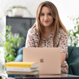 Między Myślami - Kobieta z długimi włosami siedzi przy szklanym stoliku z laptopem, obok leżą książki, w tle zielone rośliny.