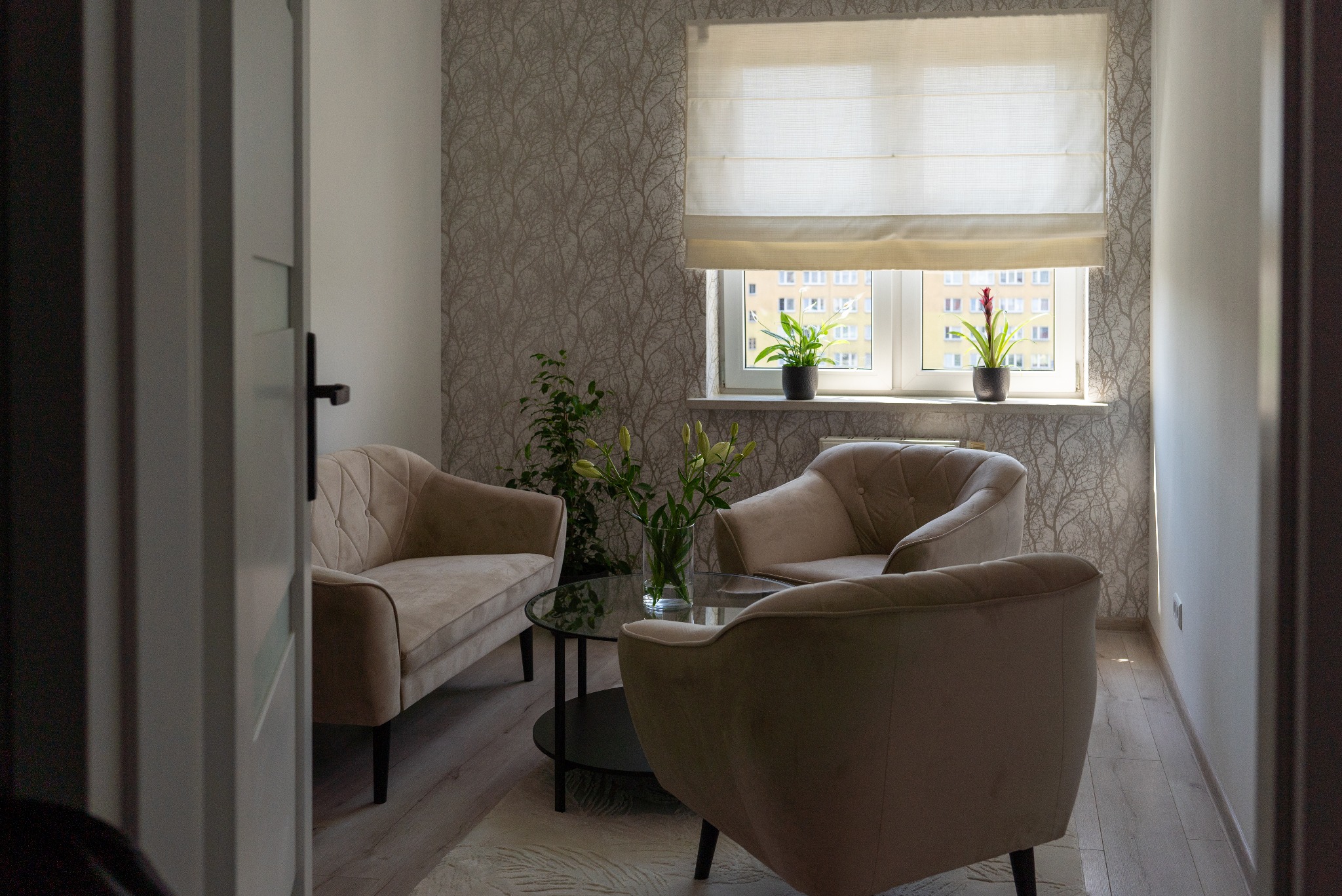 Przytulne wnętrze gabinetu psychoterapeuty z beżowymi fotelami i kanapą wokół okrągłego stolika, widoczne przez otwarte drzwi, z roślinami w doniczkach na parapecie i tapetą w motywy drzew.