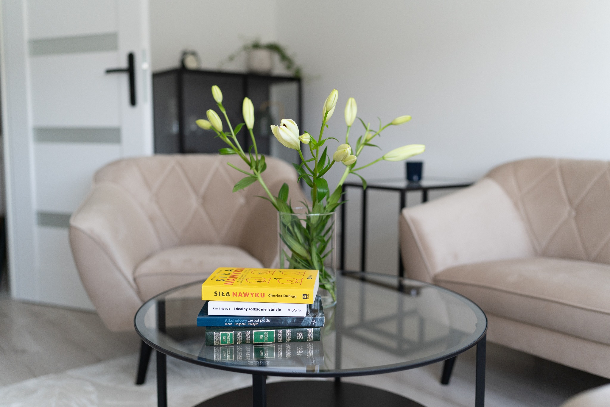 Jasne wnętrze gabinetu: na szklanym stoliku bukiet białych lilii w wazonie i stos książek, w tle dwa beżowe fotele i czarna szafka.