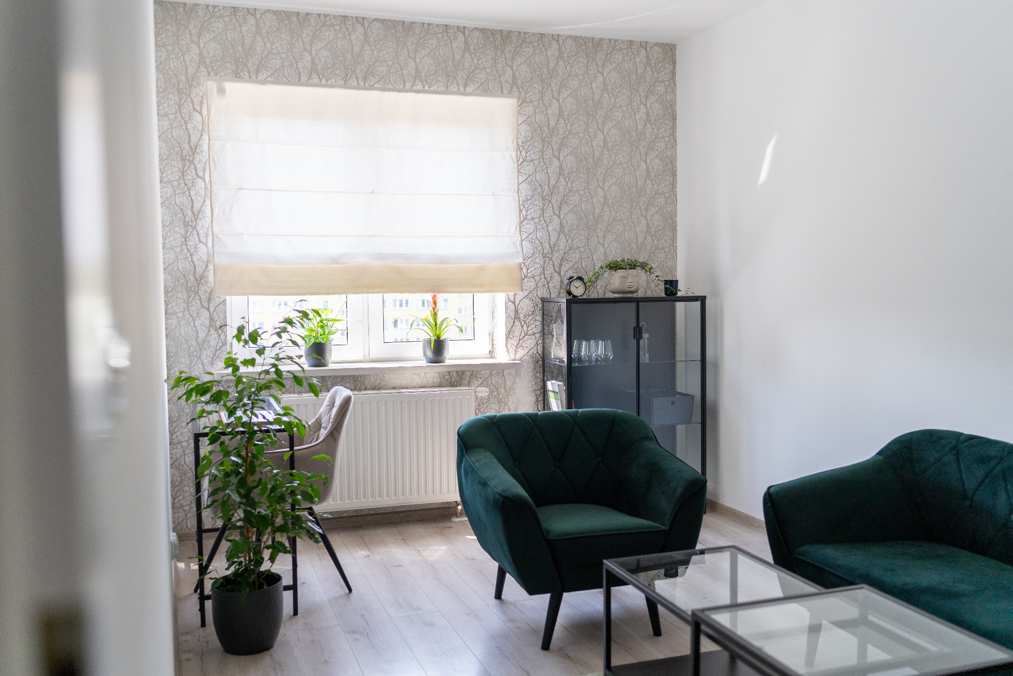 Jasne wnętrze pokoju z zielonymi fotelami, stolikiem kawowym, biurkiem z krzesłem i roślinami doniczkowymi przy oknie z roletą rzymską.