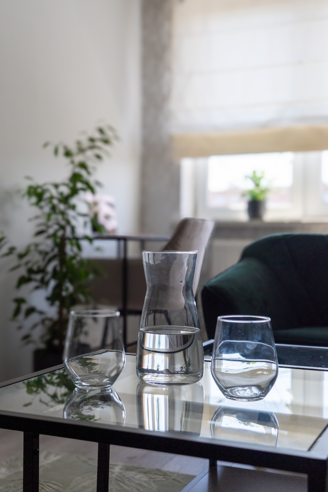 Szklany dzbanek i dwa kieliszki z wodą na szklanym stoliku kawowym, w tle zielona sofa i roślina doniczkowa, minimalistyczny wystrój wnętrza.