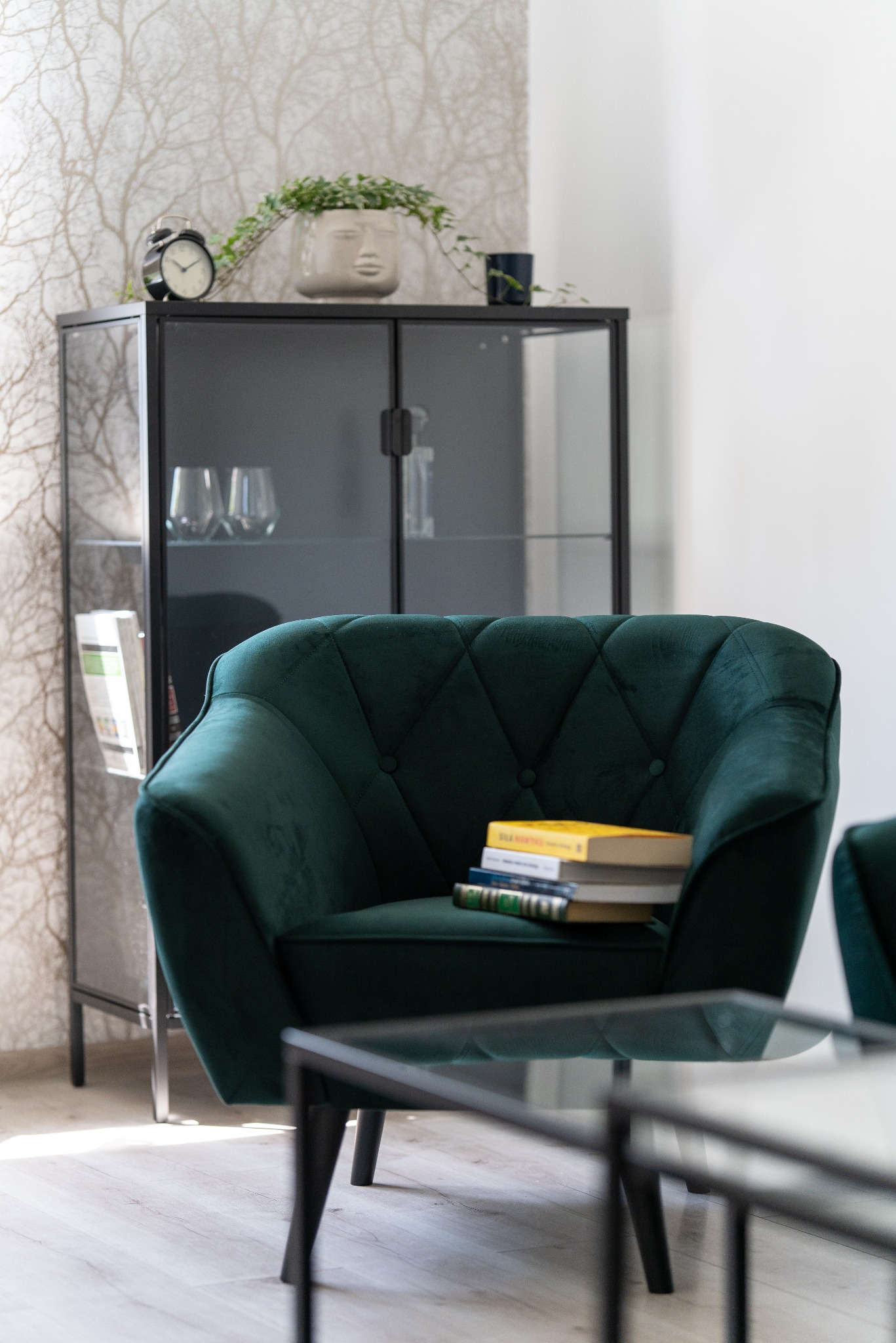 Szmaragdowy fotel z guzikami i stosem książek w eleganckim wnętrzu z przeszkloną witryną, zegarem i rośliną w doniczce z twarzą.