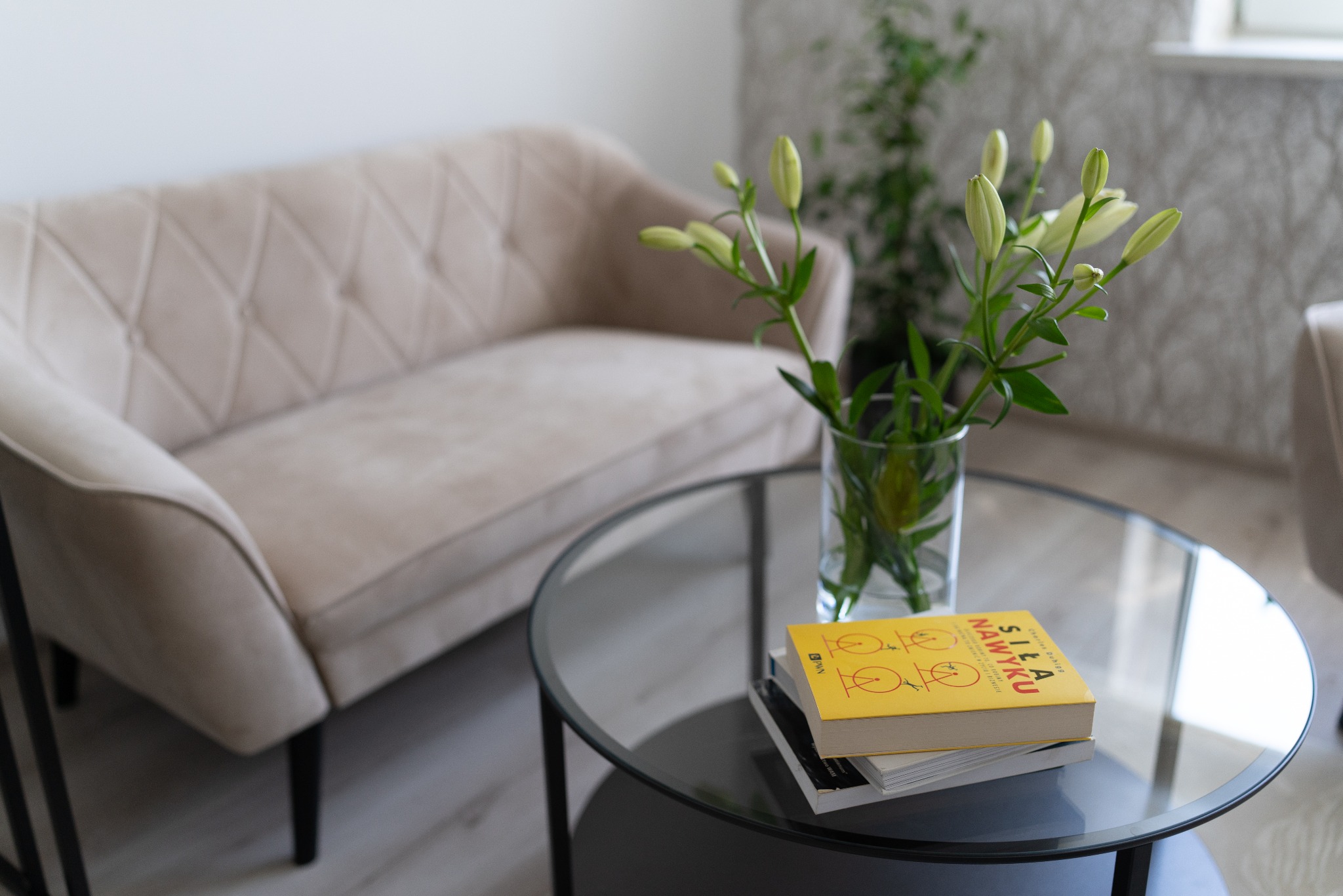 Jasny gabinet psychoterapeuty: beżowa sofa z pikowaniem, szklany stolik z wazonem lilii i książką 'Siła nawyku'.