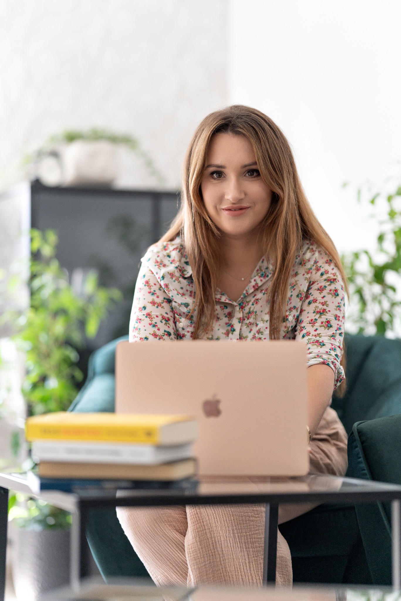 Kobieta z długimi włosami siedzi przy szklanym stoliku z laptopem, obok leżą książki, w tle zielone rośliny.