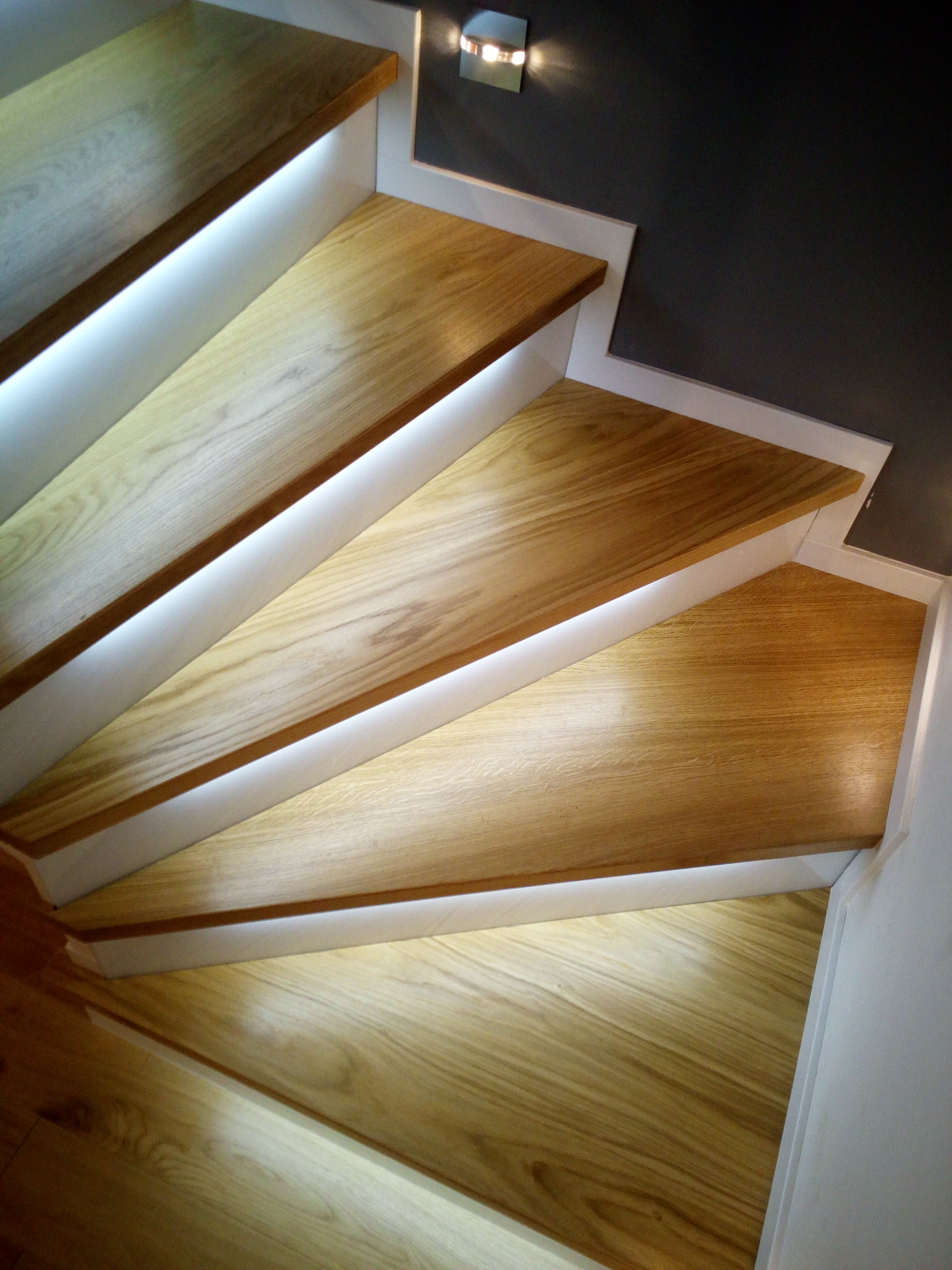 Drewniane schody z podświetleniem LED pomiędzy stopniami i białą obudową, widok z góry, na ścianie kinkiet.