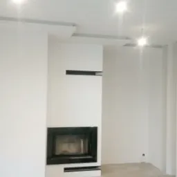 Białe, świeżo pomalowane wnętrze z czarnym kominkiem i podwieszanym sufitem z oświetleniem punktowym, widoczne ślady po taśmie malarskiej na ścianie.