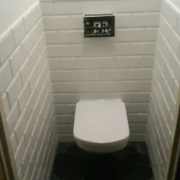 Mała toaleta z białymi, prostokątnymi płytkami na ścianach ułożonymi w cegiełkę, czarną podłogą i podwieszaną muszlą klozetową z czarnym panelem spłukującym.