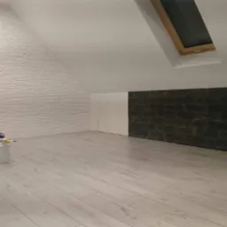 Poddasze po remoncie: biała cegła dekoracyjna na skosie, ciemna okładzina na ścianie, jasne panele podłogowe, wiadro z farbą i narzędziami malarskimi przy ścianie z cegły.