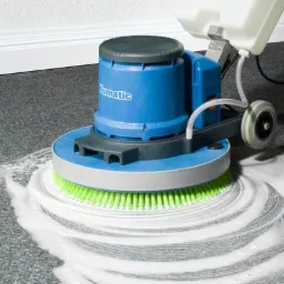 Profesjonalne czyszczenie tekstylnej wykładziny podłogowej za pomocą szorowarki z zieloną szczotką i zbiornikiem na wodę, widoczne ślady detergentu.