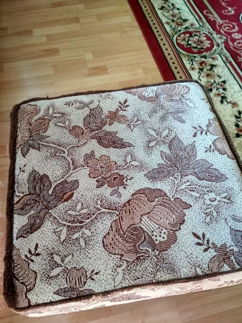 Kwadratowa pufa z brązowym, kwiatowym wzorem na beżowym tle, obita brązowym futerkiem, leżąca na drewnianej podłodze obok dywanu w kwiaty.