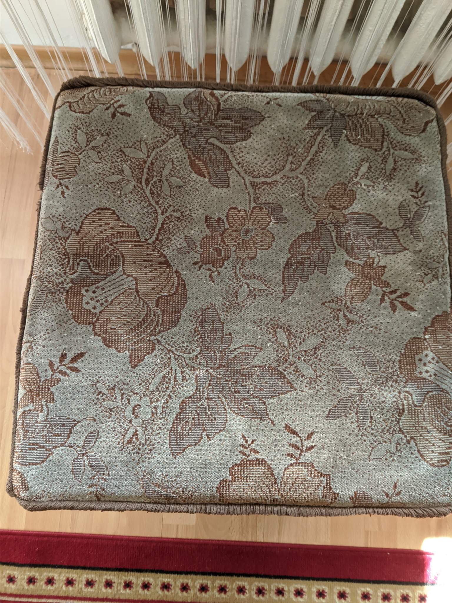 Tapicerowany stołek z brązowo-beżowym wzorem kwiatowym, widok z góry, z frędzlami na krawędzi, na tle drewnianej podłogi i fragmentu czerwonego dywanu.