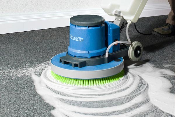 Profesjonalne czyszczenie tekstylnej wykładziny podłogowej za pomocą szorowarki z zieloną szczotką i zbiornikiem na wodę, widoczne ślady detergentu.