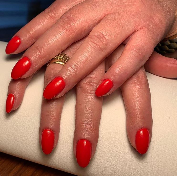 Czerwone, błyszczące paznokcie w Studzienicach, owalny kształt, złoty pierścionek na palcu wskazującym, kremowe tło.