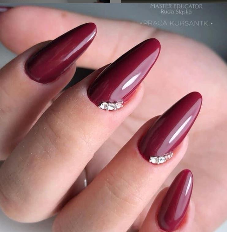 Burgundowe, błyszczące paznokcie w kształcie migdała, zdobione drobnymi kryształkami przy nasadzie, na tle dłoni.
