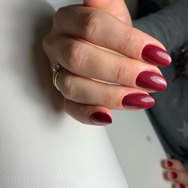 Dłoń z pomalowanymi na burgundowo owalnymi paznokciami, na serdecznym palcu złoty pierścionek z oczkiem, na jasnym tle.