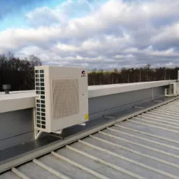 Kaskada zewnętrznych jednostek klimatyzacyjnych marki Gree zamontowanych na dachu budynku przemysłowego, widok z perspektywy dachu, pochmurne niebo w tle.