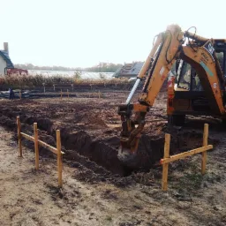 Żółta koparka w trakcie wykonywania wykopu pod fundamenty, widoczne drewniane kołki wyznaczające obszar prac, w tle zabudowania i szklarnia.