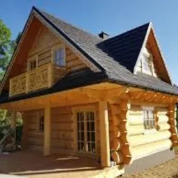 Nowoczesny, parterowy dom z bali z poddaszem użytkowym, czarnym dachem i drewnianym balkonem z ażurową balustradą, widoczny z perspektywy ogrodu w słoneczny dzień.