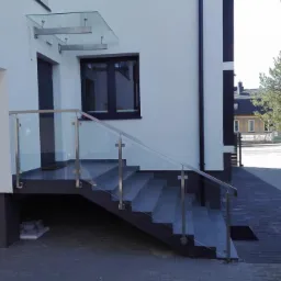 Zewnętrzne schody z szarych płytek z balustradą ze stali nierdzewnej i szkła, prowadzące do wejścia domu z szklanym zadaszeniem.