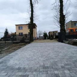 Świeżo ułożona, szara kostka brukowa na podjeździe posesji, widoczne elementy ogrodzenia w trakcie montażu i palety z kostką w tle.