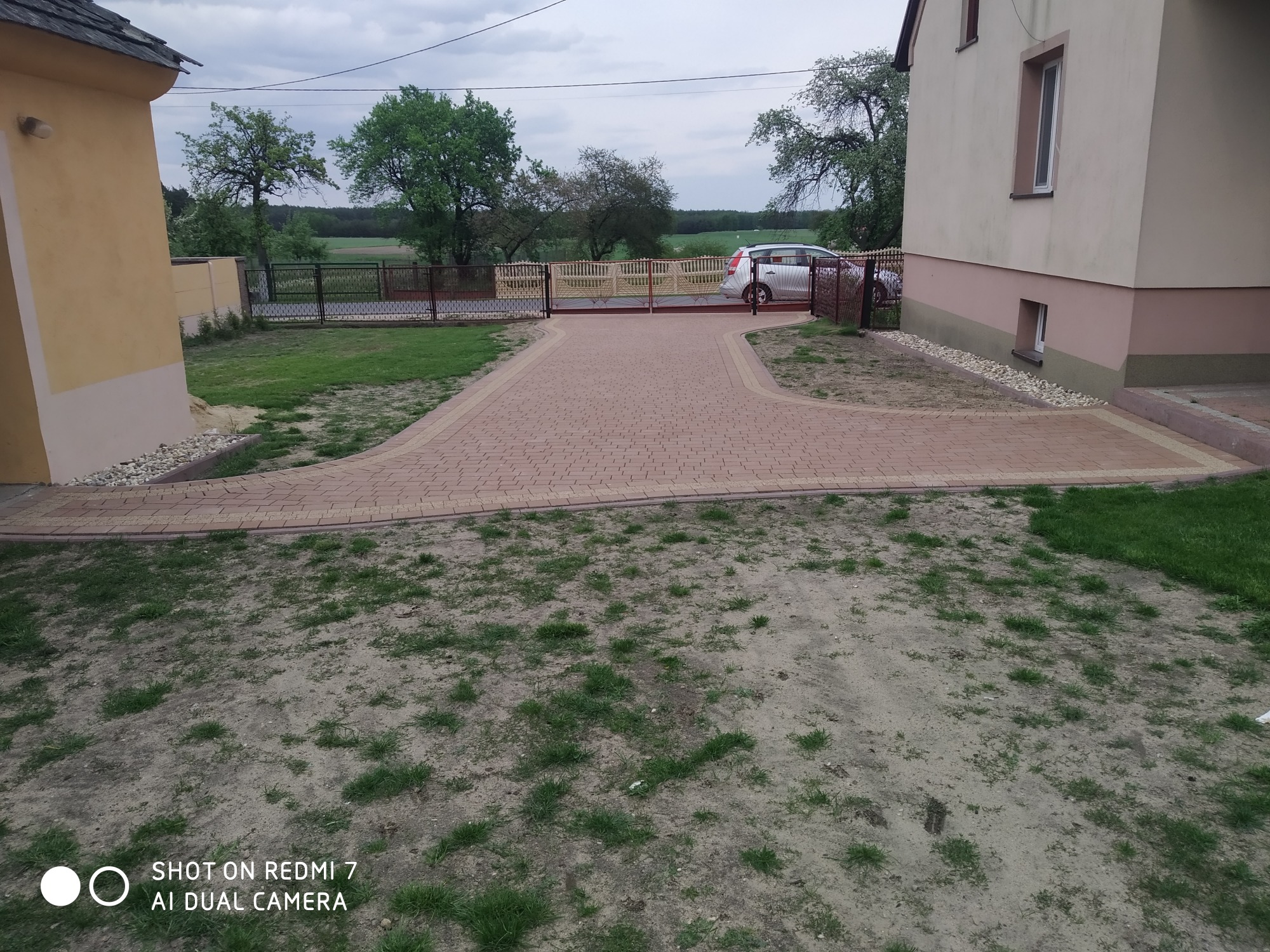 Ułożona, czerwona kostka brukowa tworząca podjazd do posesji z bramą wjazdową, widok na dom i ogród w tle.