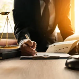 Adwokat w garniturze sporządza dokumenty na biurku z wagą Temidy, kodeksem i młotkiem sędziowskim w słonecznym biurze