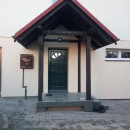 Zadaszone wejście do domu z ciemnozielonymi drzwiami, drewnianymi słupami i czerwoną dachówką, obok skrzynka pocztowa, na schodach buty i talerz z resztkami jedzenia, na chodniku śmieci.