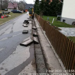 Mini koparka przy krawężniku, obok wykopana bruzda pod instalacje, porozrzucane płyty chodnikowe, drewniany płot w tle.