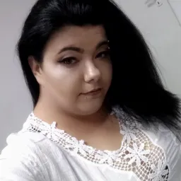 Portret kobiety z ciemnymi włosami, z delikatnym makijażem oczu i piercingiem na brodzie oraz nad brwią, ubrana w białą, koronkową bluzkę.